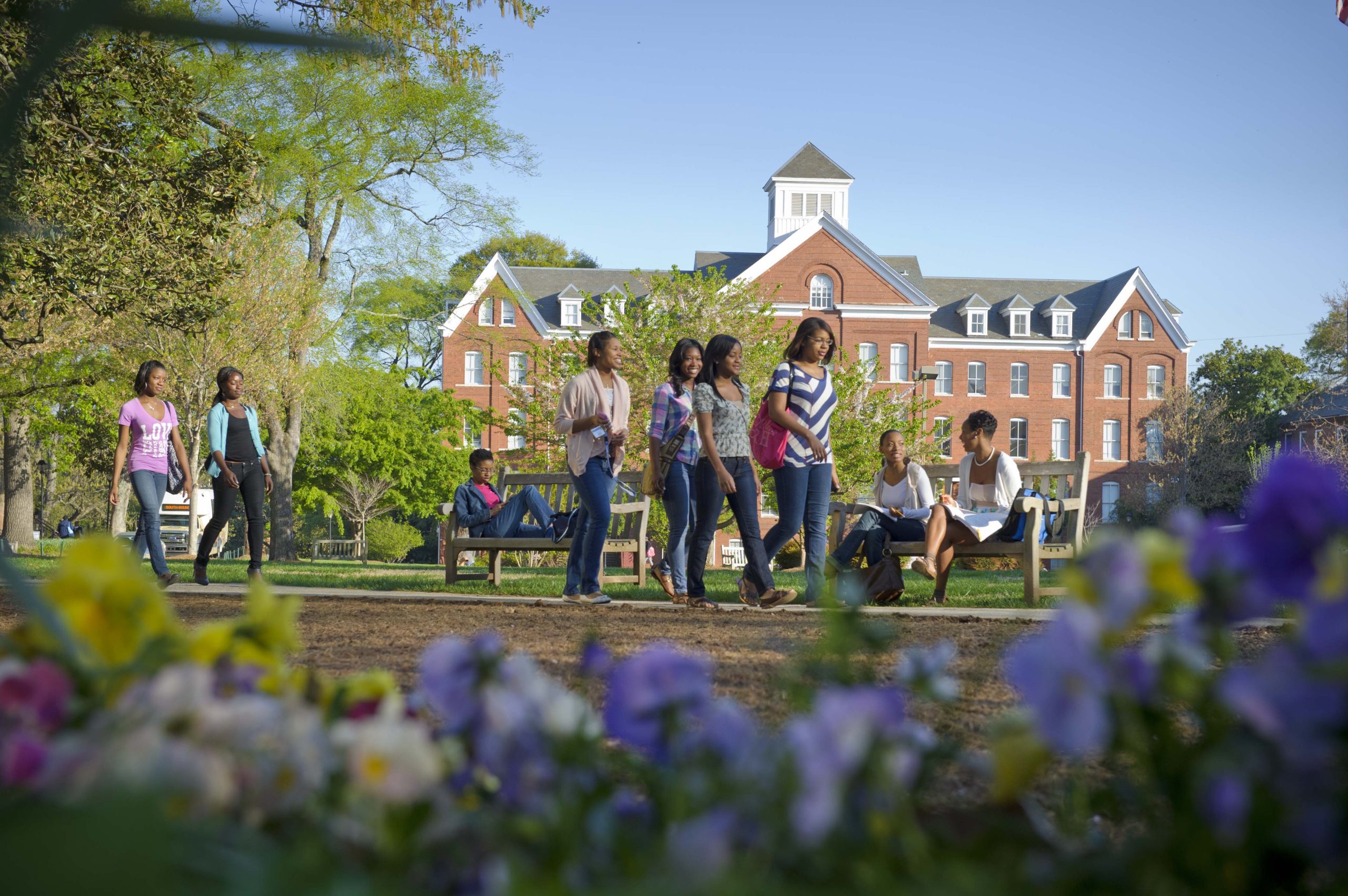 The Spemlan College Campus with students walking to class and sitting on benches. There is a school building in the background and green trees on the left 