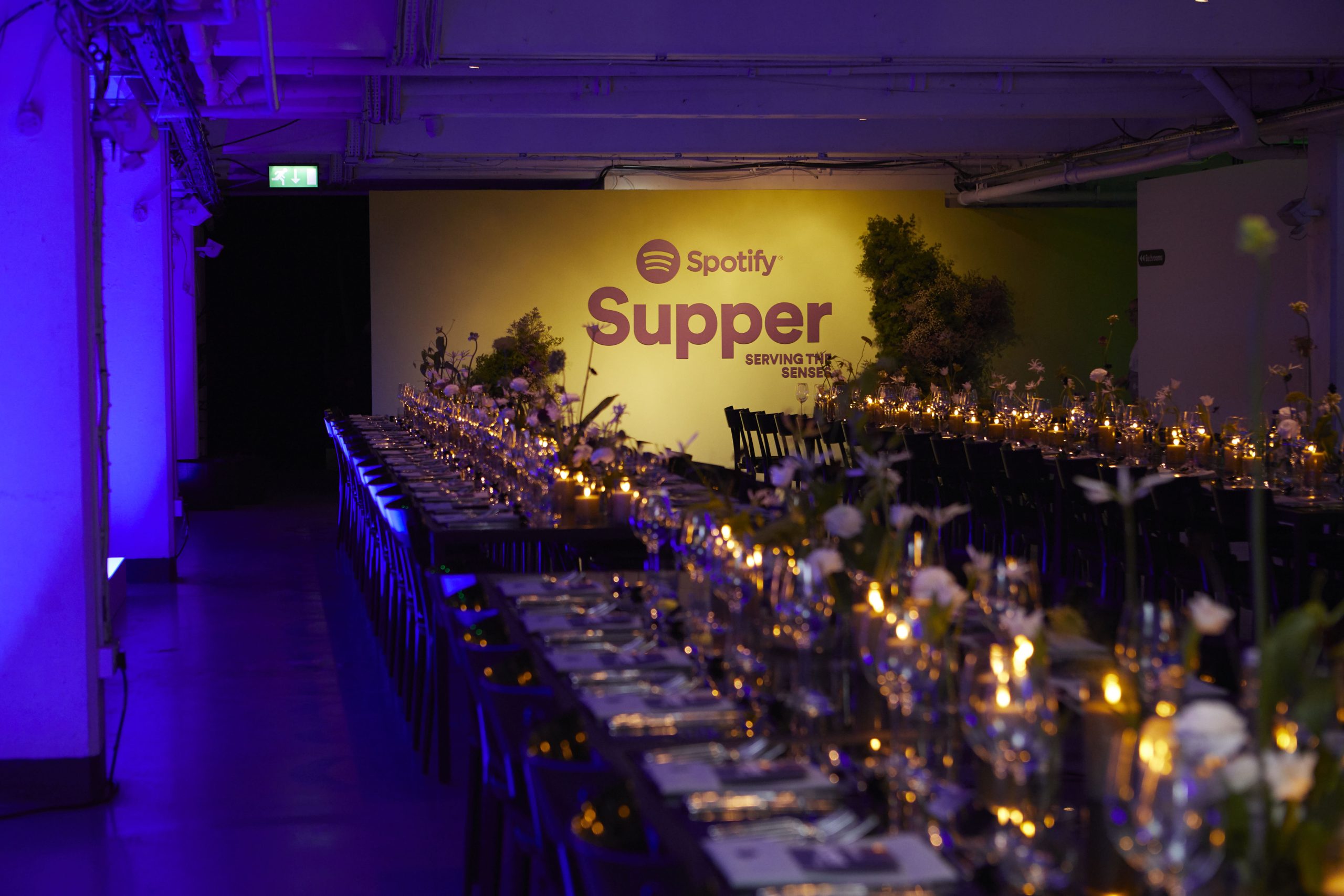 long tables set with plates, flowers, and candles for the spotify supper. the back drop has a spotify supper sign 