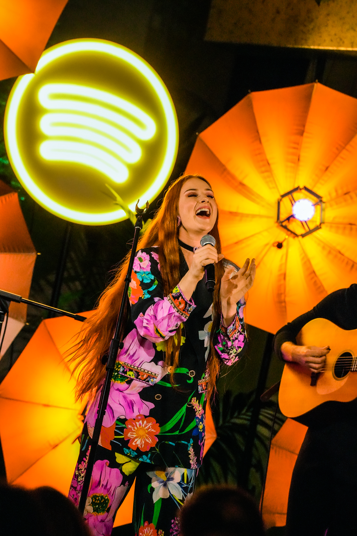 Singer, Vera Blue, holding a microphone and singing at a Spotify supper. She is wearing a colorful pantsuit with a pink floral pattern