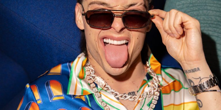 peso pluma holds his sunglasses over his eyes while sticking his tongue out