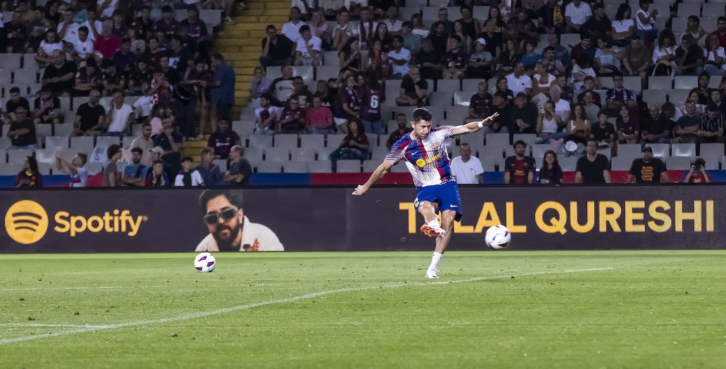 FCB soccer player kicking a ball on the field. A spotify sign is in the background