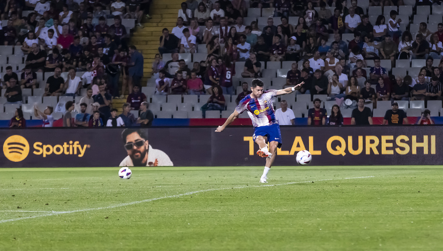 FCB soccer player kicking a ball on the field. A spotify sign is in the background