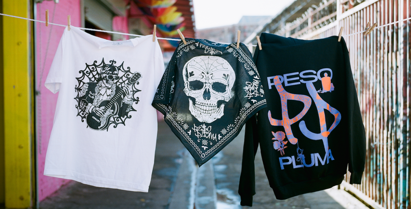 Peso Pluma's merch hanging on a clothing line outside. There is a white tshirt with a skull, a black scarf with a white skull, and a black sweatshirt with Peso Pluma's name