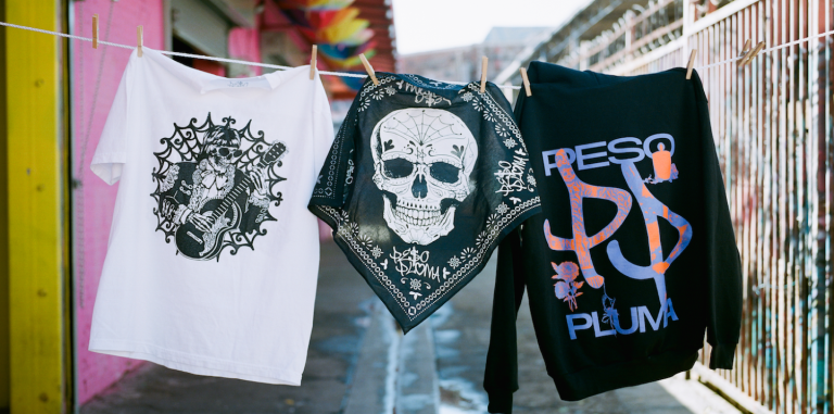 Peso Pluma's merch hanging on a clothing line outside. There is a white tshirt with a skull, a black scarf with a white skull, and a black sweatshirt with Peso Pluma's name