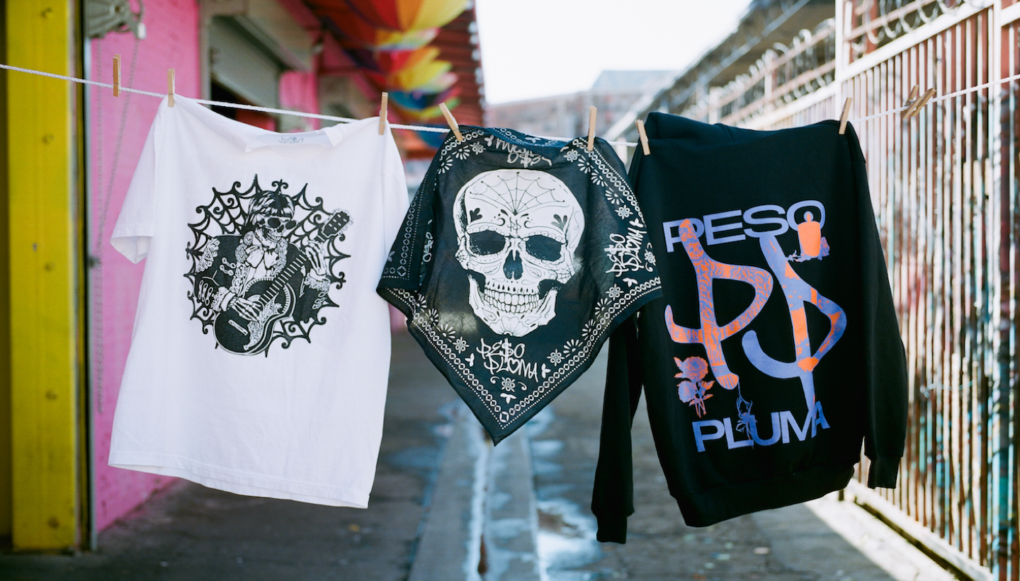 Peso Pluma's merch hanging on a clothing line outside. There is a white tshirt with a skull, a black scarf with a white skull, and a black sweatshirt with Peso Pluma's name