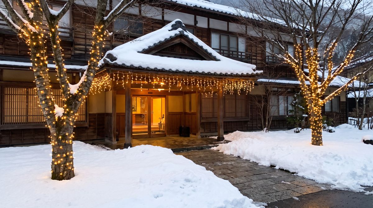 雪の夕方の温泉旅館の玄関。軒先に電球色のつららライトを下げ、両脇の木の幹に電球色のストレートライトを巻いて点灯している
