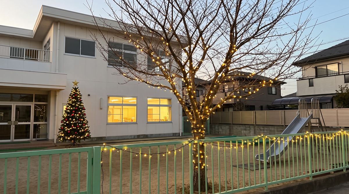 夕暮れの幼稚園の園庭。フェンスと園庭の木に電球色のストレートライトを付け、園舎の入口の脇にクリスマスツリーを置いて点灯している