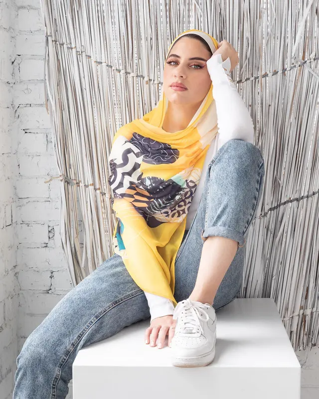 Casual outfit with a yellow printed hijab, white long-sleeve top, light-wash rolled jeans and white trainers, model seated against a textured backdrop.