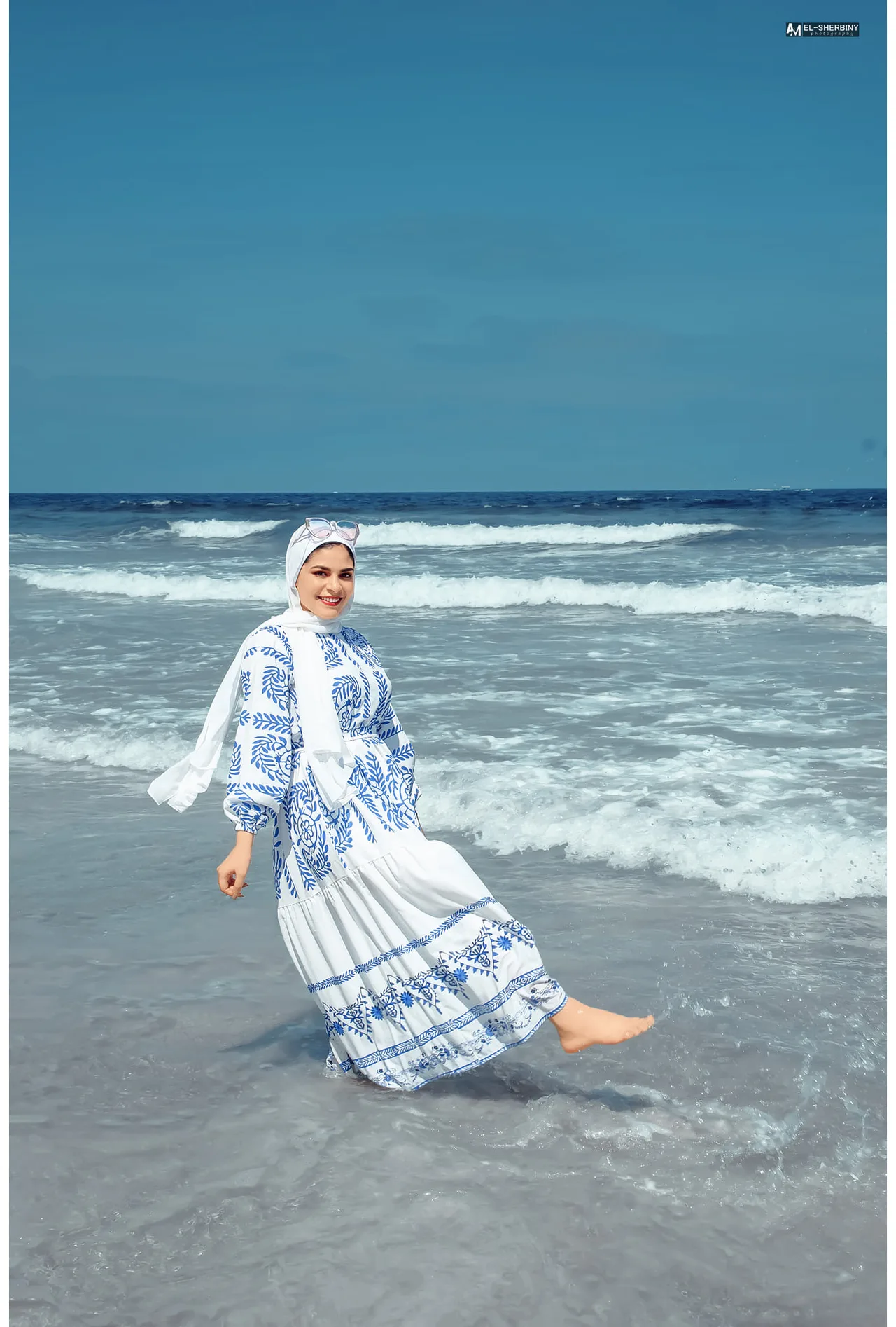 White maxi dress with blue botanical print and white hijab worn by a woman standing barefoot at the seashore, sunglasses pushed up over the hijab.