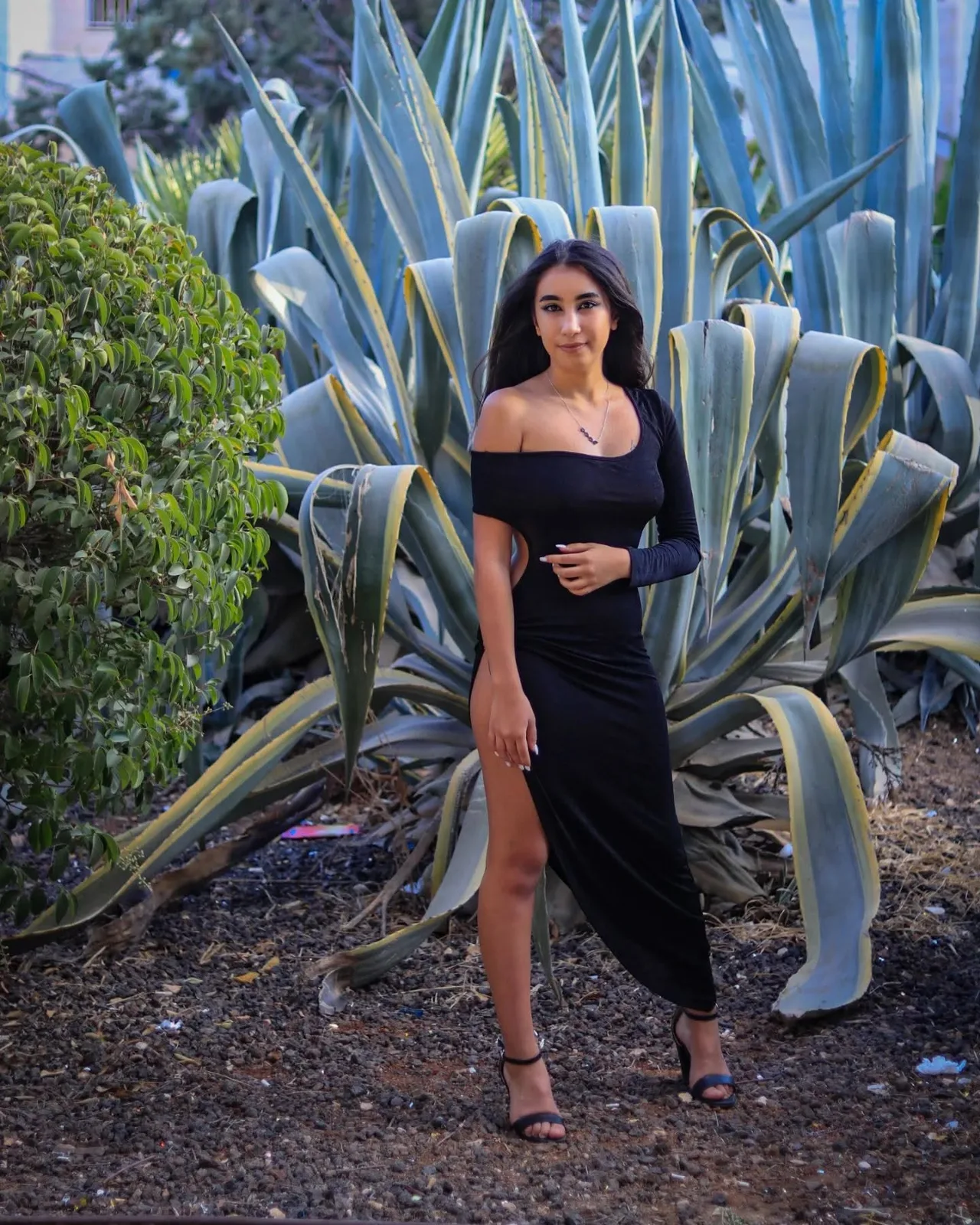 Black one-sleeve midi dress with side cut-out and high thigh slit worn with black heeled sandals and a silver necklace.