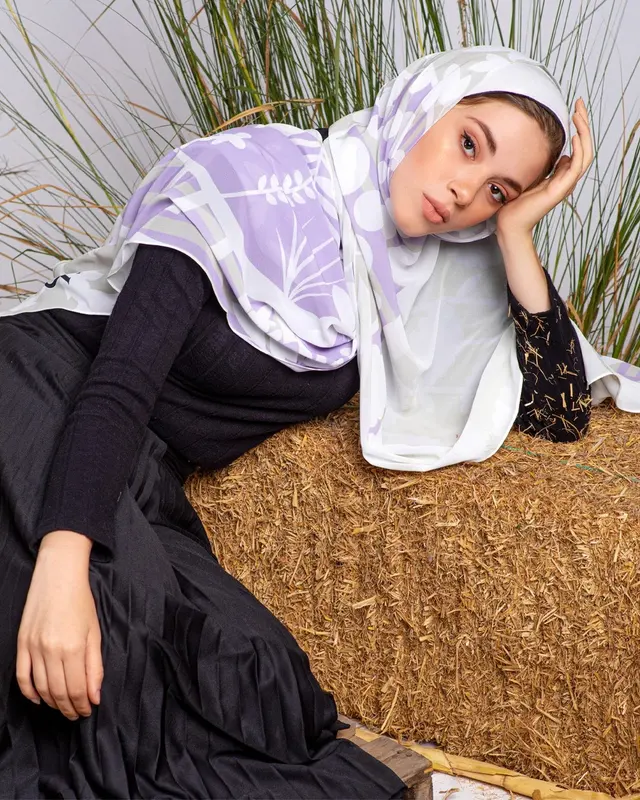 Layered outfit with a lilac and white printed hijab draped over a black ribbed long-sleeve top and a black pleated maxi skirt, model reclining on a straw bale.