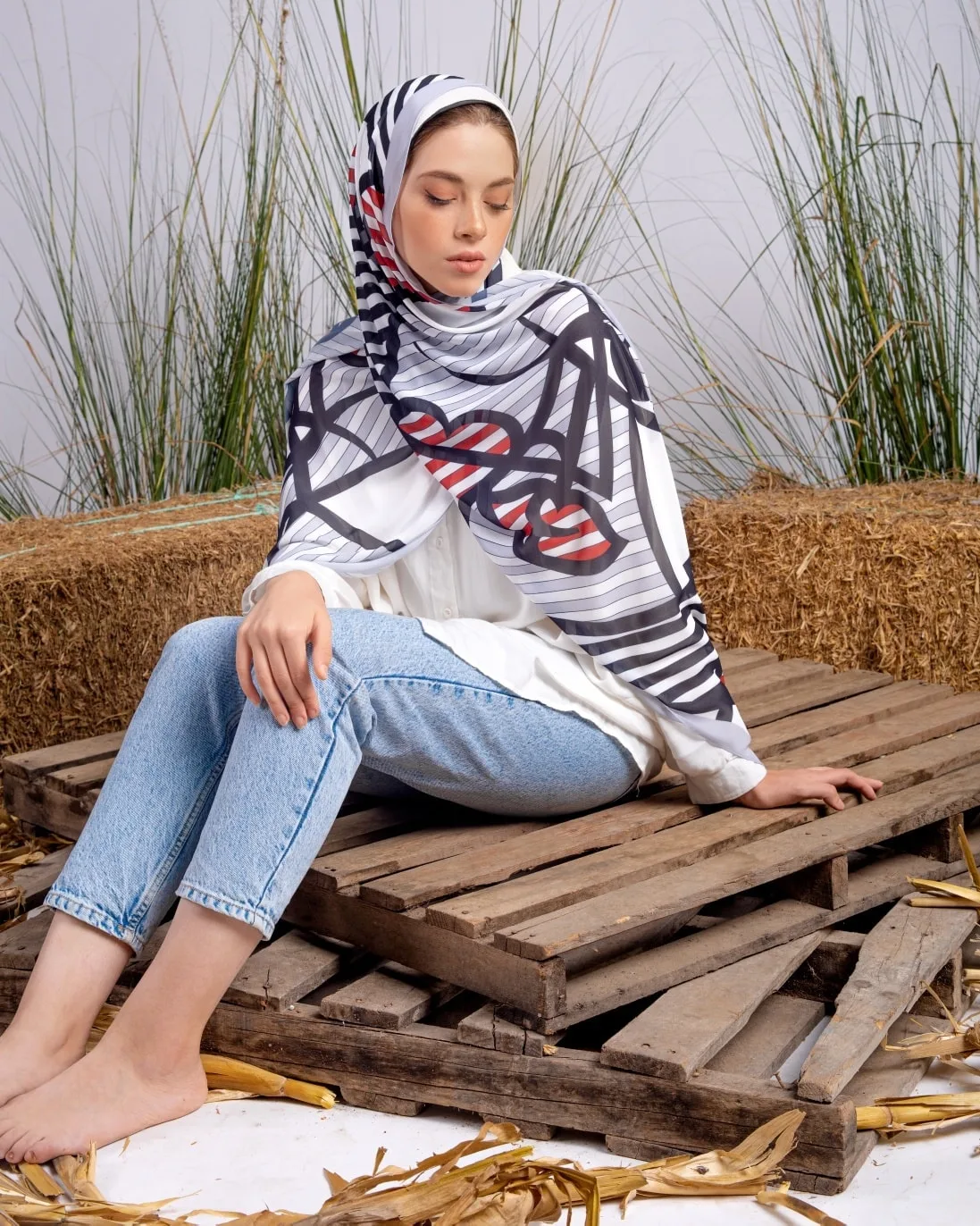 Layered look with a white printed hijab, white button-down shirt and light wash straight jeans, model seated on wooden pallets.