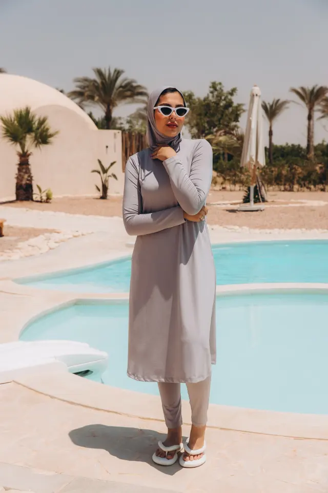 Light grey long-sleeve midi dress layered over matching slim trousers with a coordinating hijab, white cat-eye sunglasses and white padded sandals by a pool.