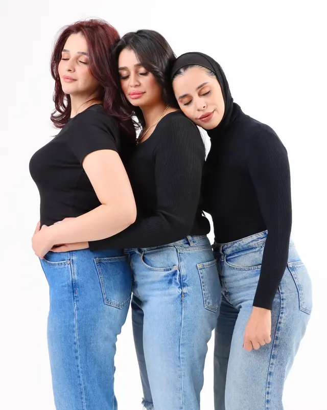 Three women wearing black tops and light blue jeans standing side by side, left in a short-sleeve crew t-shirt, centre in a ribbed scoop-neck sweater with gold chain, right in a ribbed turtleneck and black headscarf.