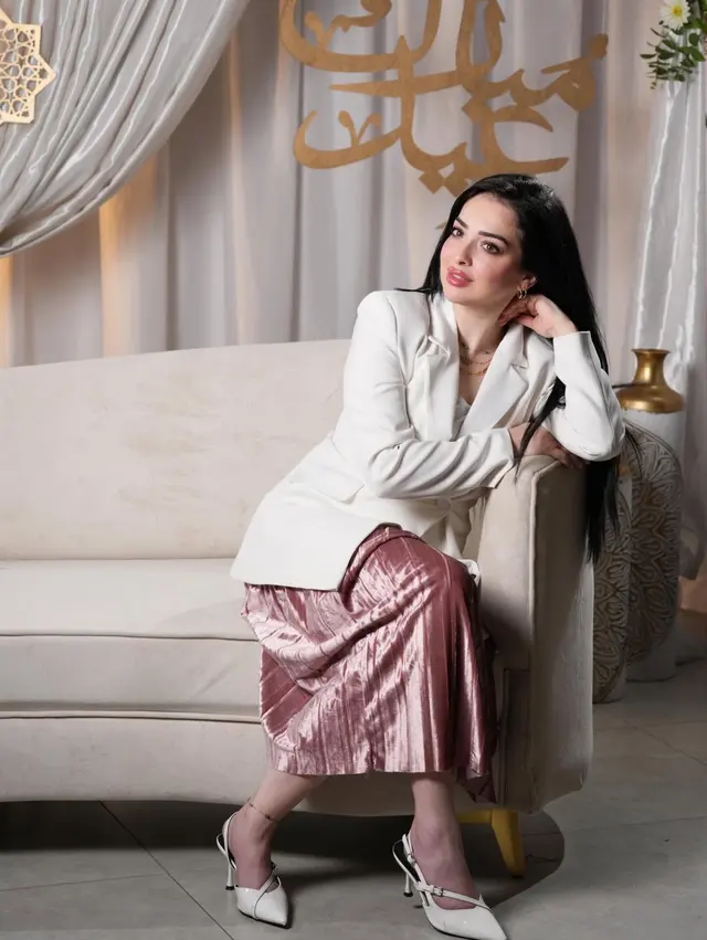 White tailored blazer paired with a pleated pink midi skirt, white slingback heels and layered gold necklaces, woman seated on a pale sofa.