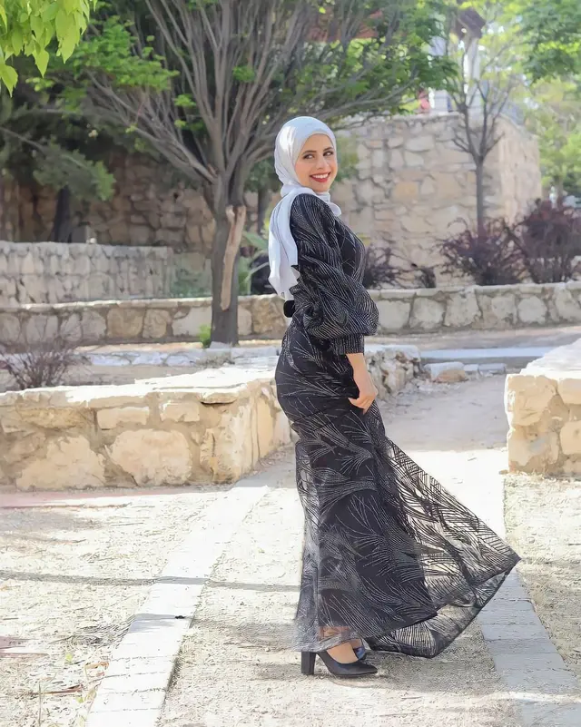 Black patterned maxi dress with long puffed sleeves worn with a white hijab and black heels, model standing outdoors by stone walls.