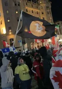 A group of protesters gathering outdoors. One is holding a Bitcoin flag, several others hold Canadian flags.