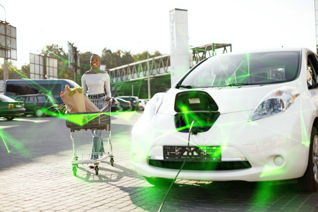 Woman pushing trolley through car park next to electric vehicle being charged with green plexus overlayed