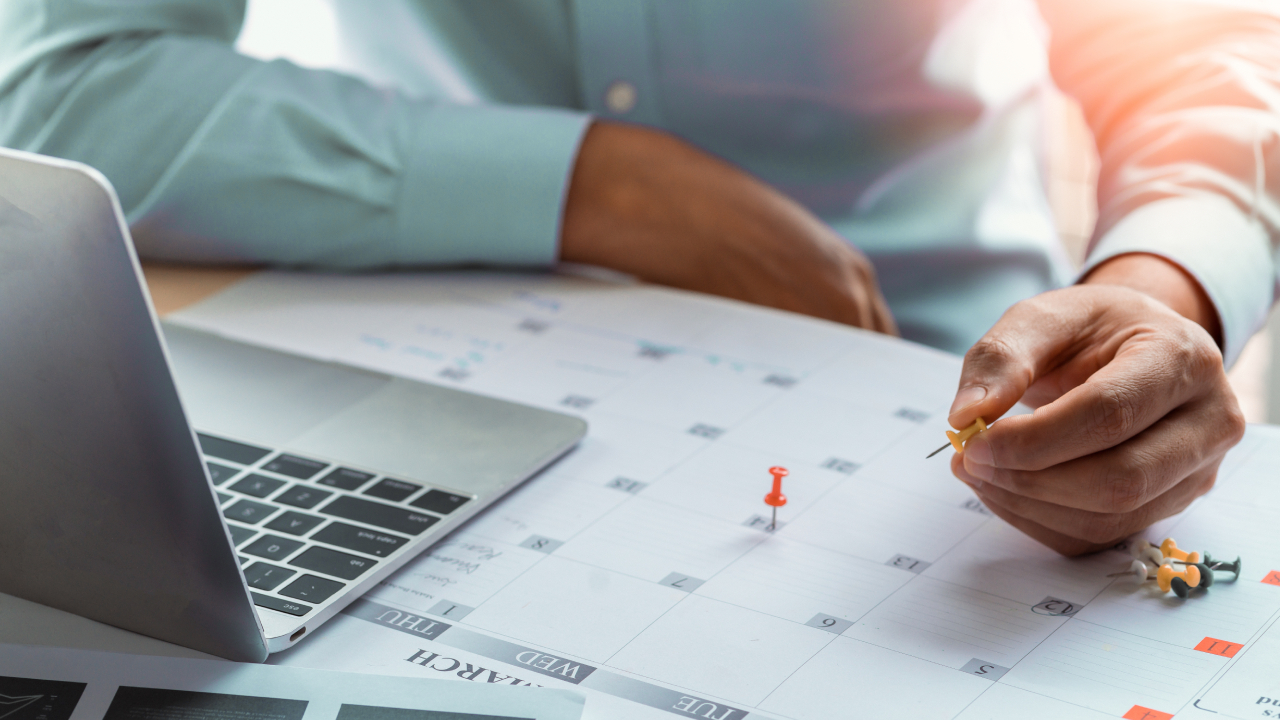 Man marking travel dates on a calendar with push pins while planning a school holiday trip on his laptop.