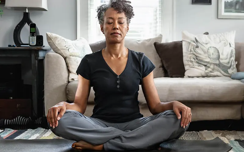 A woman sitting on a yoga mat peacefully meditating in her living room.