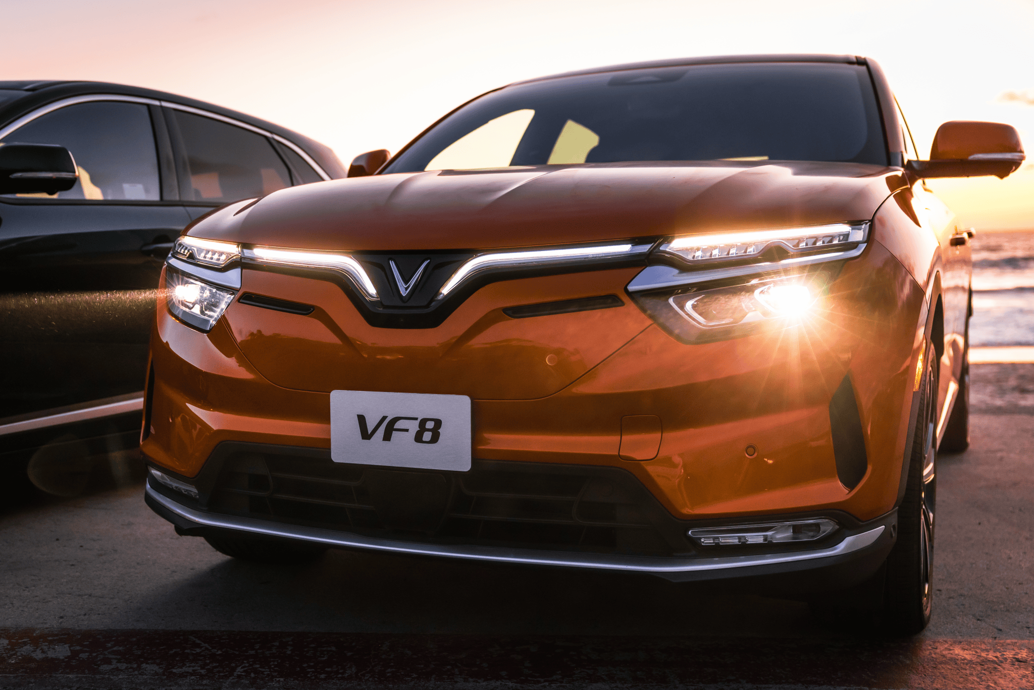 An orange VinFast VF 8, a California EV, is parked on a sandy beach along the coast at golden hour.