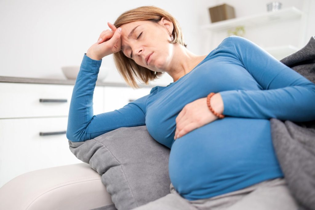 Pregnant tired exhausted woman with stomach and head issues at home on a couch, being sick.