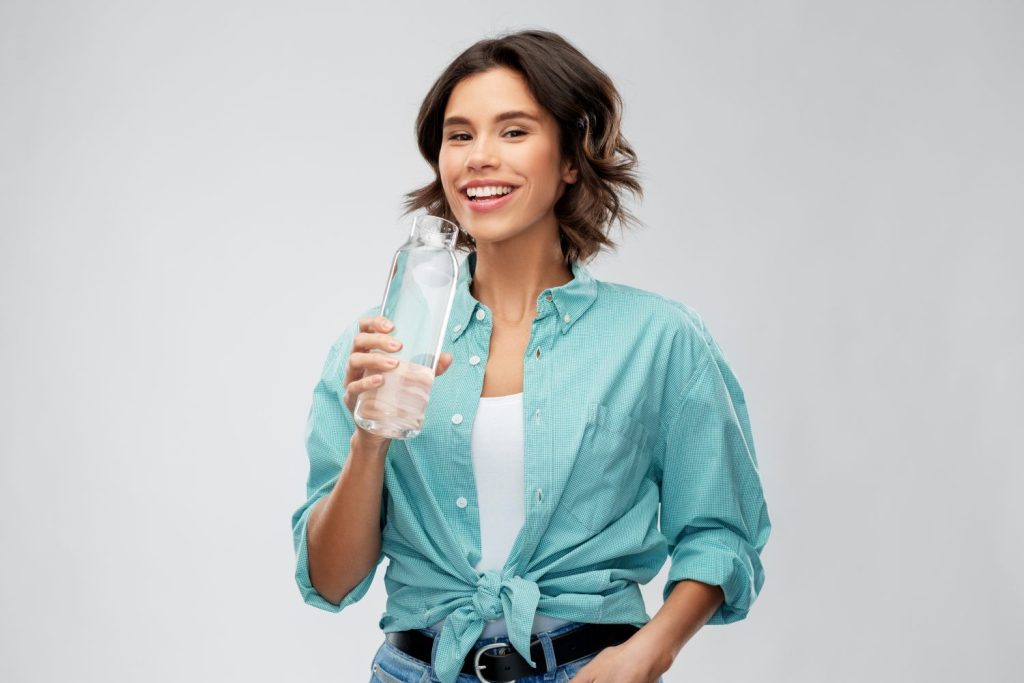 people concept - portrait of happy smiling young woman in turquoise shirt drinking water from reusable glass bottle grey background