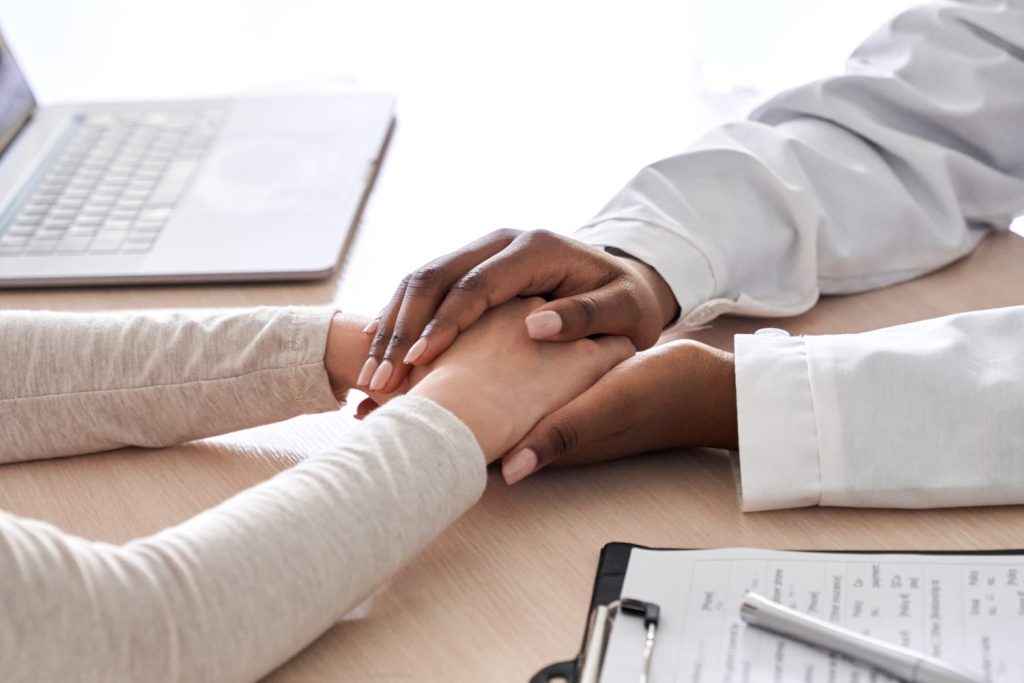 African female doctor holding hand supporting caucasian woman patient. Kind ethnic professional physician give empathy concept encourage reassure infertile patient at medical visit, close up view.