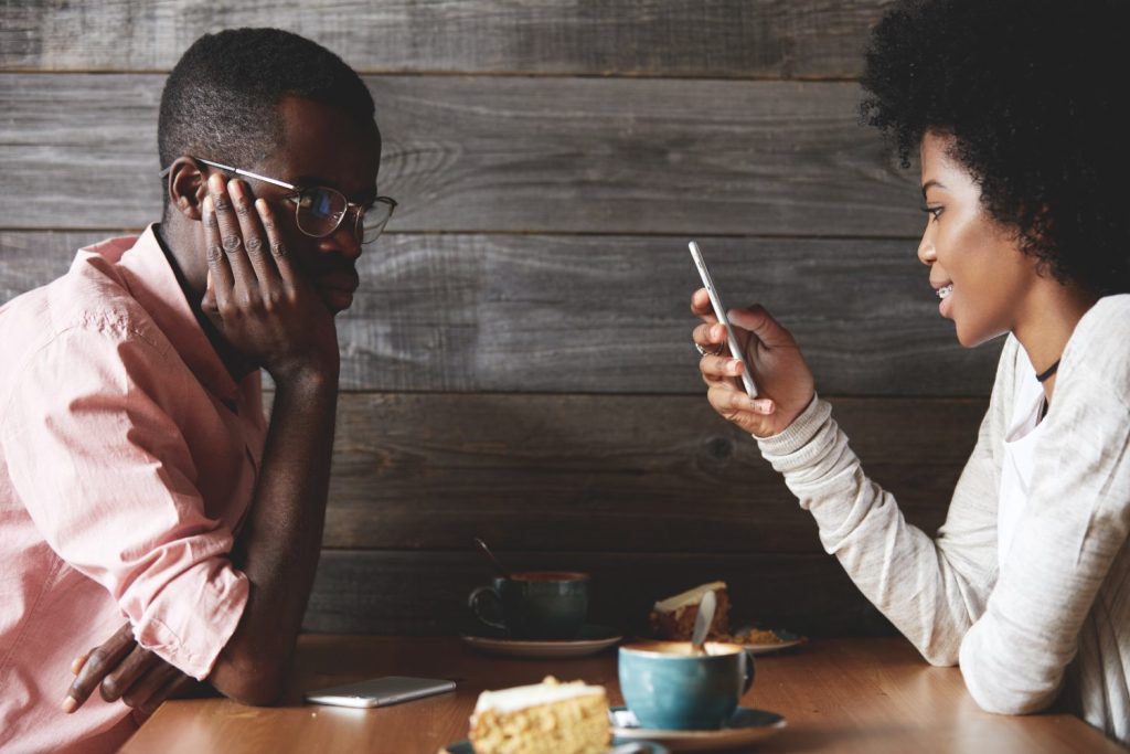 Young gloomy and bored African man getting angry while his cheerful dark skinned girlfriend chatting with her friends on social networks using mobile phone, instead of talking to him during the date