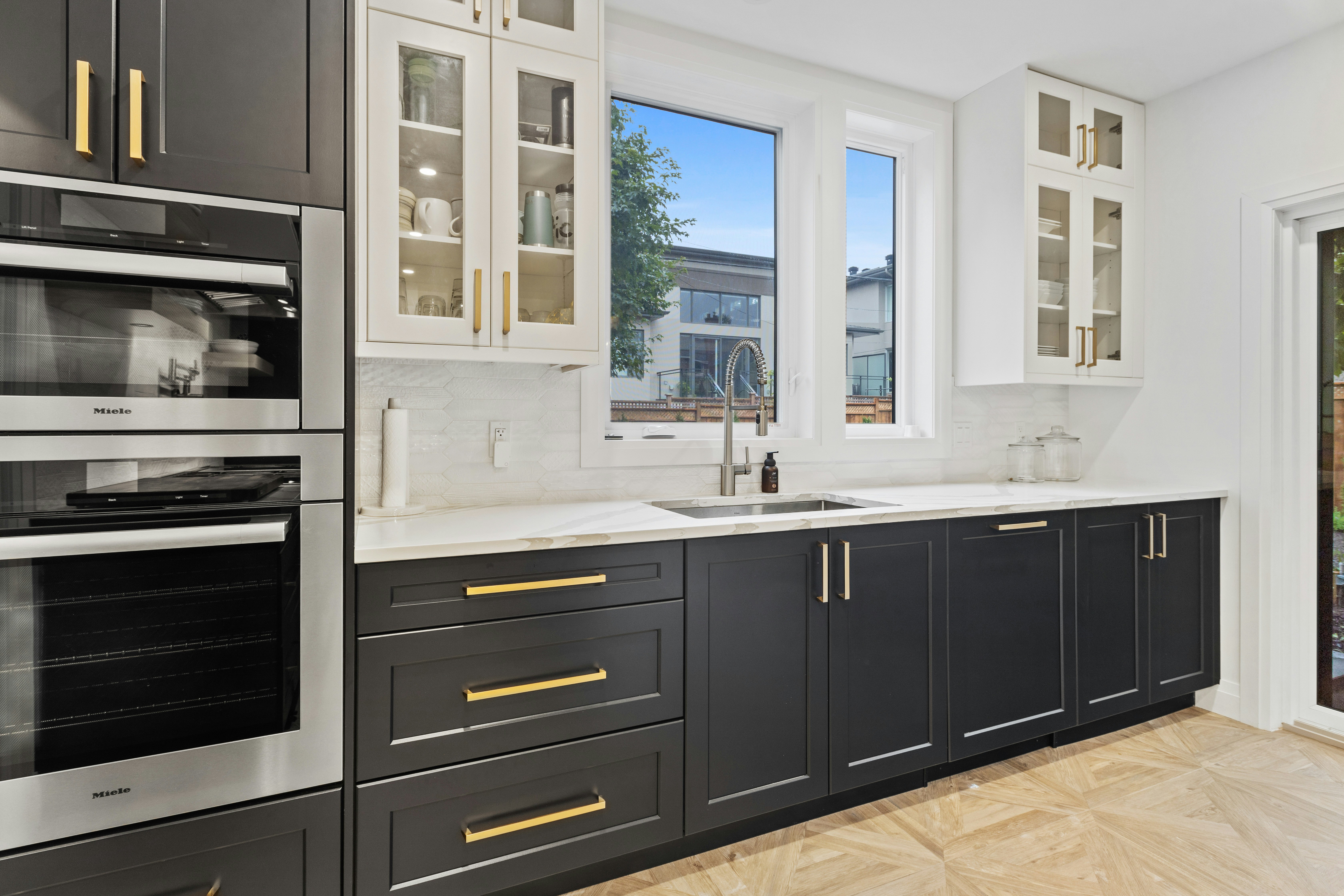 Modern kitchen featuring sleek black cabinetry with gold hardware, built-in Miele ovens, white countertops, and large windows letting in natural light.