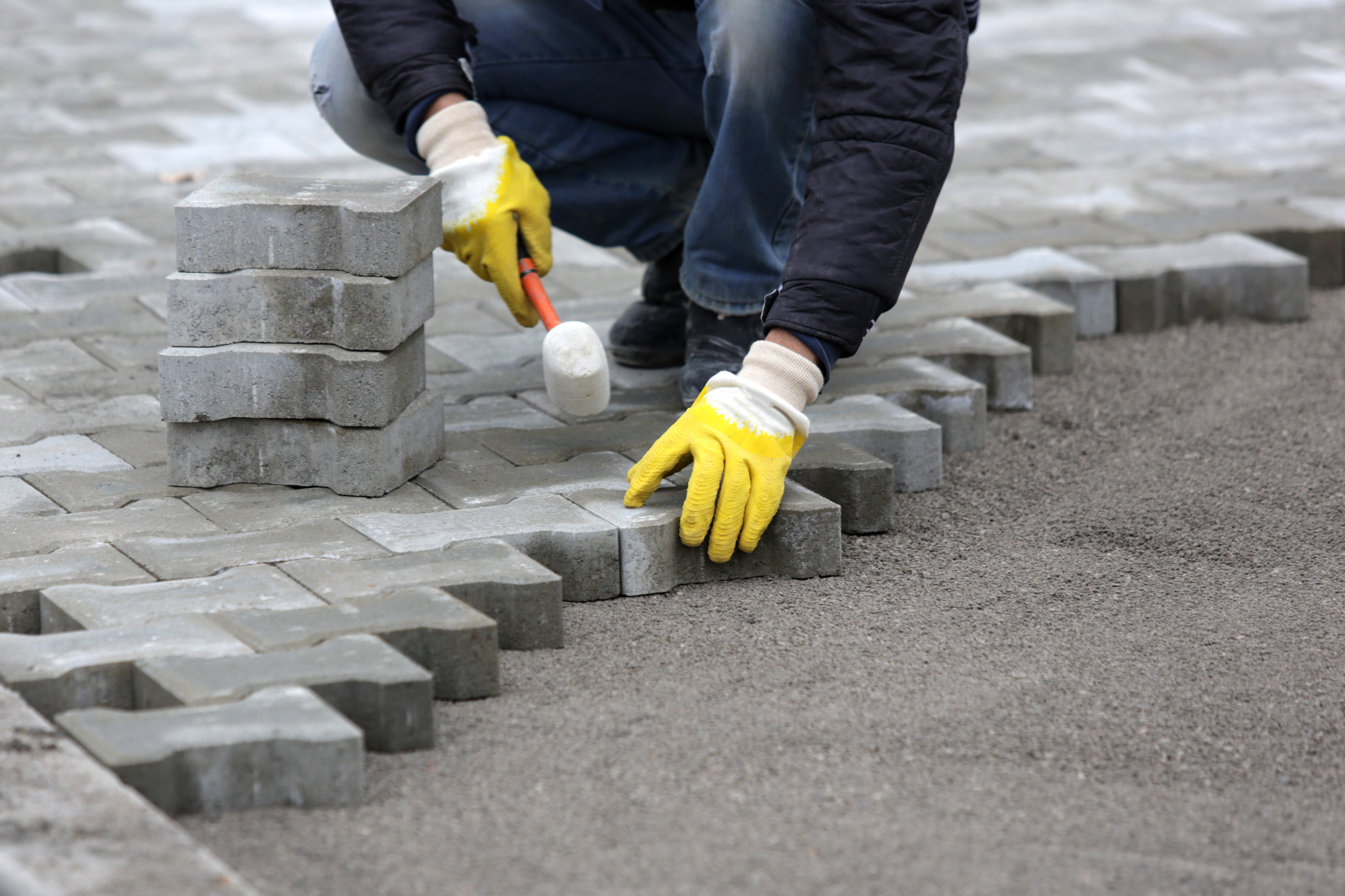 A worker kneels, placing gray paving stones in a neat pattern on a sandy surface.