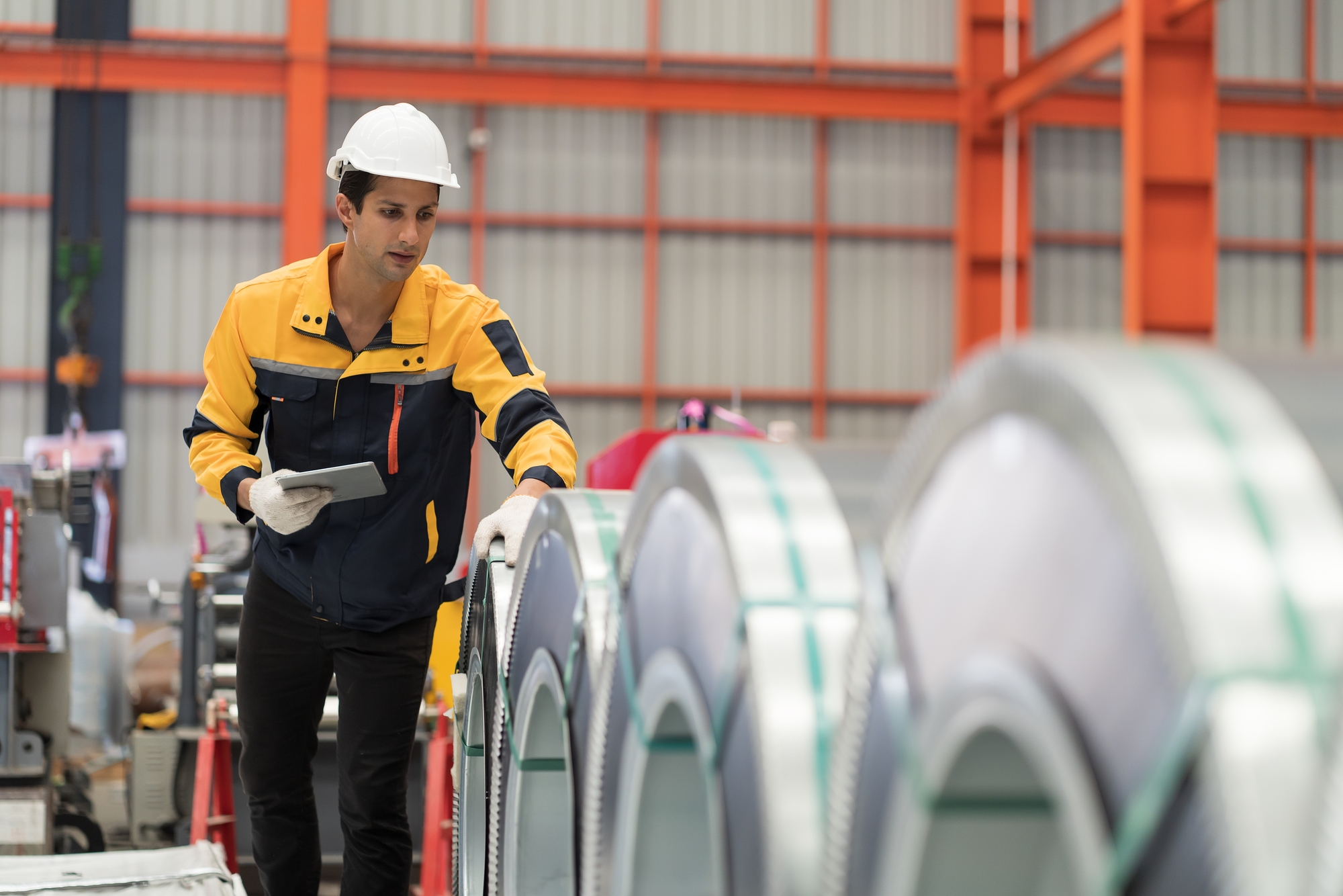 A man in a yellow shirt and hard hat stands beside large rolls of steel in a construction setting.