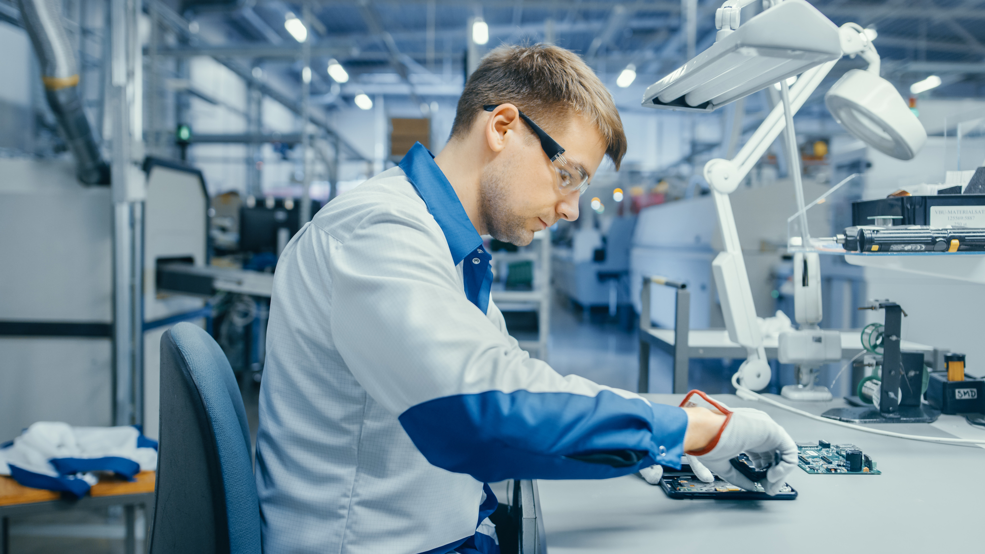 A man focused on a computer while working in a factory setting, surrounded by machinery and equipment.