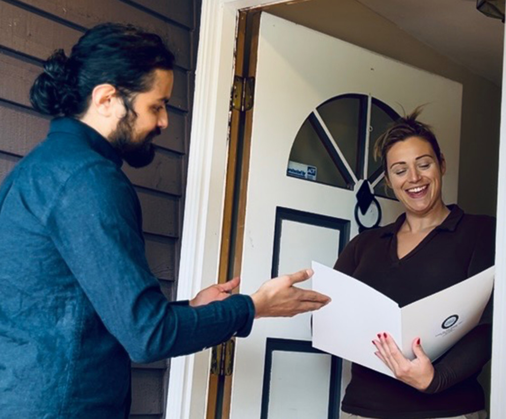 A person in a blue shirt extends their hand while a woman in a brown top holds a document at a doorway.