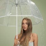 woman with long blonde hair holding a clear umbrella on a green background while rain falls down on her