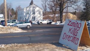 Hand painted sign next to a road that says, "zoning kills dreams," across the road from a town hall with cars in the parking lot.