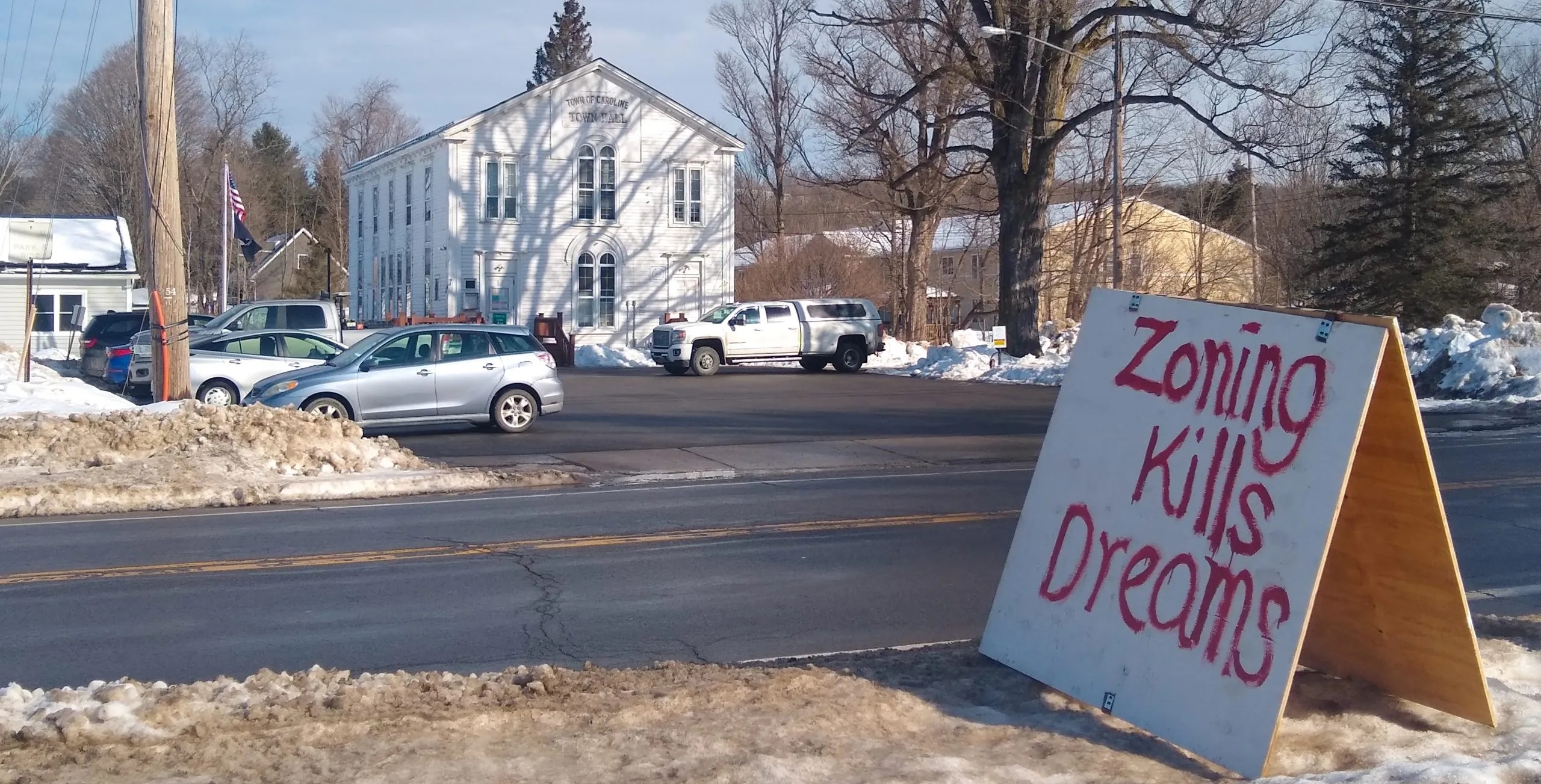 Hand painted sign next to a road that says, "zoning kills dreams," across the road from a town hall with cars in the parking lot.