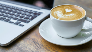 A wooden desk with a laptop keyboard and a latte in a white mug on a white saucer.