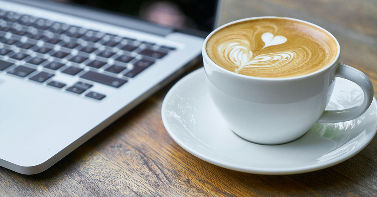 A wooden desk with a laptop keyboard and a latte in a white mug on a white saucer.