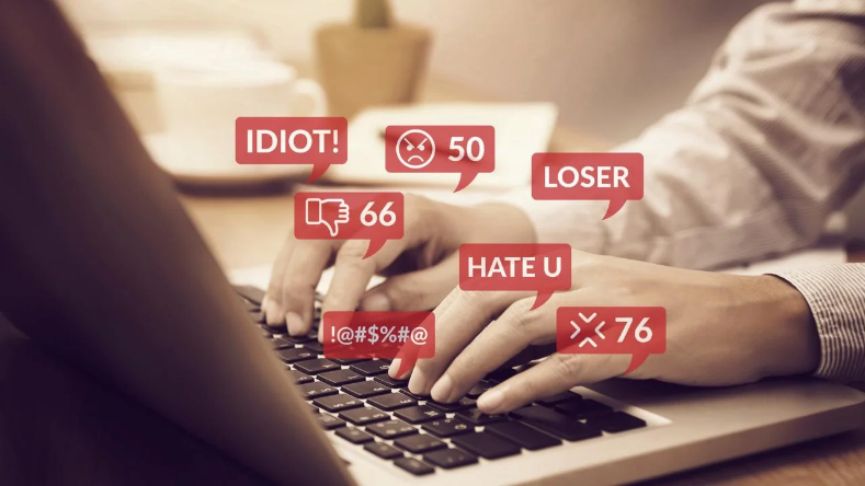 A close-up of hands typing on a laptop surrounded by red bubbles with negative messages such as "hate u" surrounding the hands and keyboard.