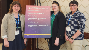 A group of 3 people pose for a photo next to a sign for a local government conference.