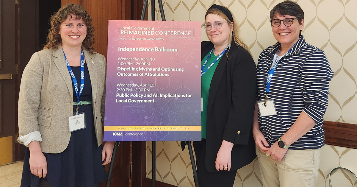 A group of 3 people pose for a photo next to a sign for a local government conference.