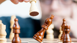 A close-up of a chessboard showing a hand above the board using a piece to knock another piece from the board.