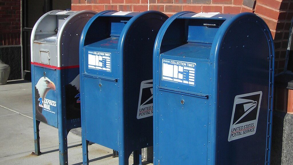 A row of 3 blue U.S. Postal Service mailboxes stand next to a brick wall.