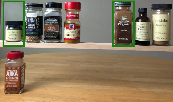 A row of 7 spice jars sit on a wooden shelf. The dill weed and paprika are outlined in green rectangles. A jar of ajika sits on a wooden countertop below.