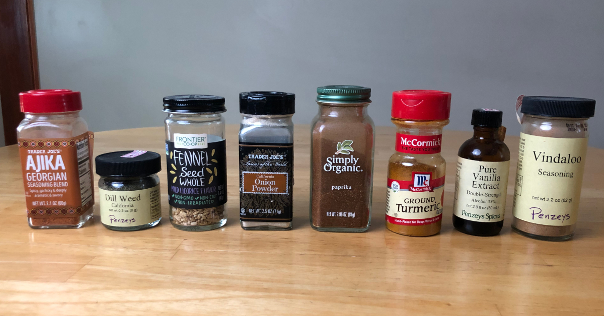 A row of 8 spice jars on a wooden countertop. The spices include ajika, dill weed, fennel seed, onion powder, paprika, turmeric, vanilla extract, and vindaloo seasoning.