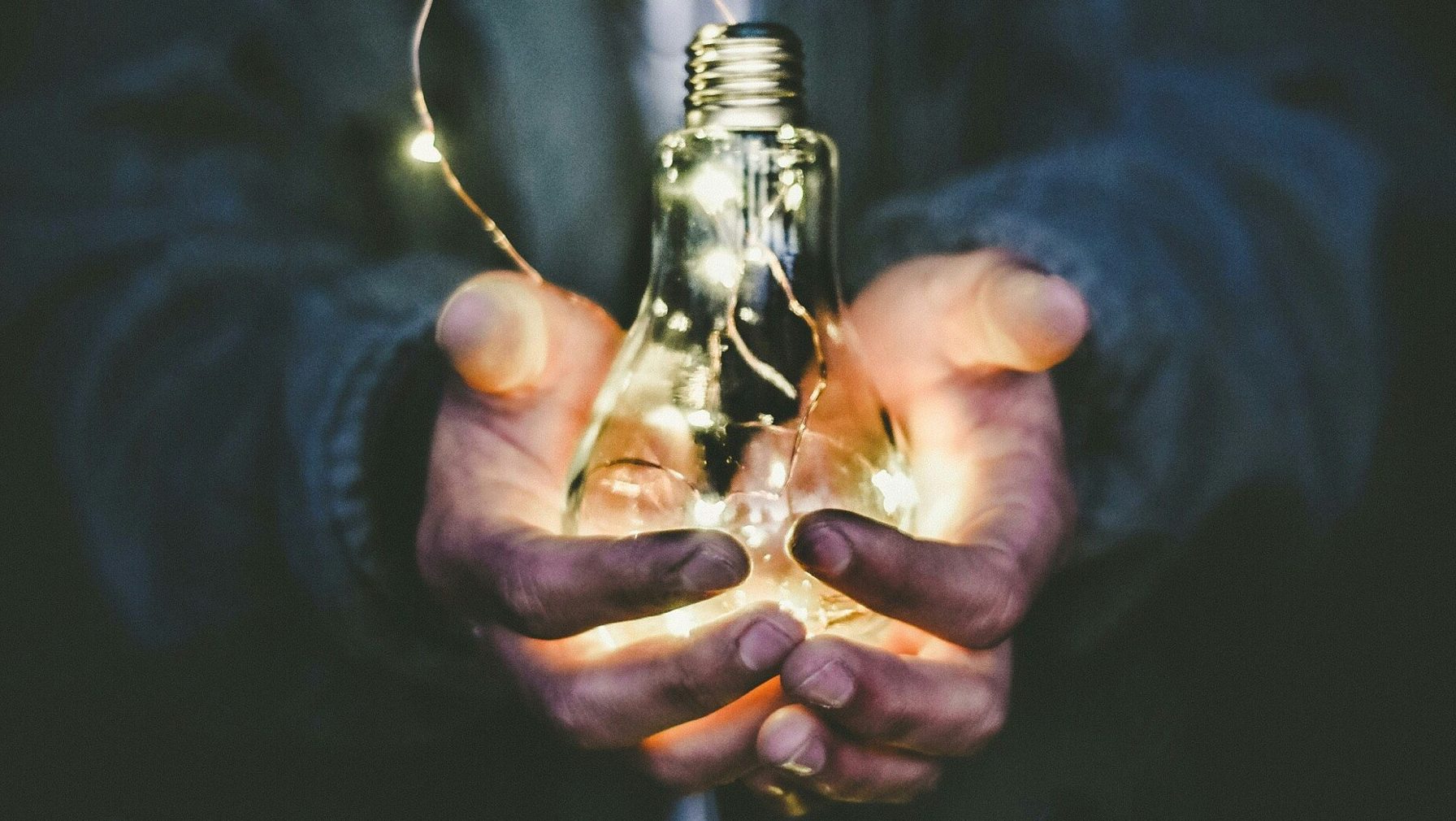 A pair of hands hold a glowing incandescent lightbulb.