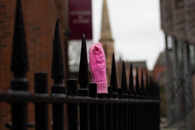 A pink, knit mitten covering a spike on a black, wrought-iron fence, with buildings in the background out of focus.