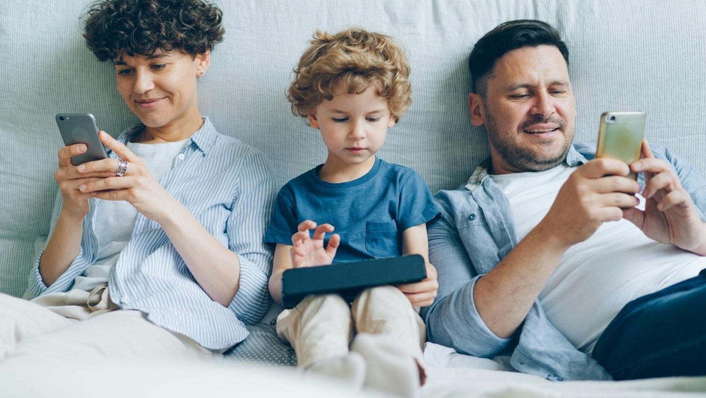 Two children and a man sit together while using smart devices. Photo by Vitaly Gariev on Unsplash.