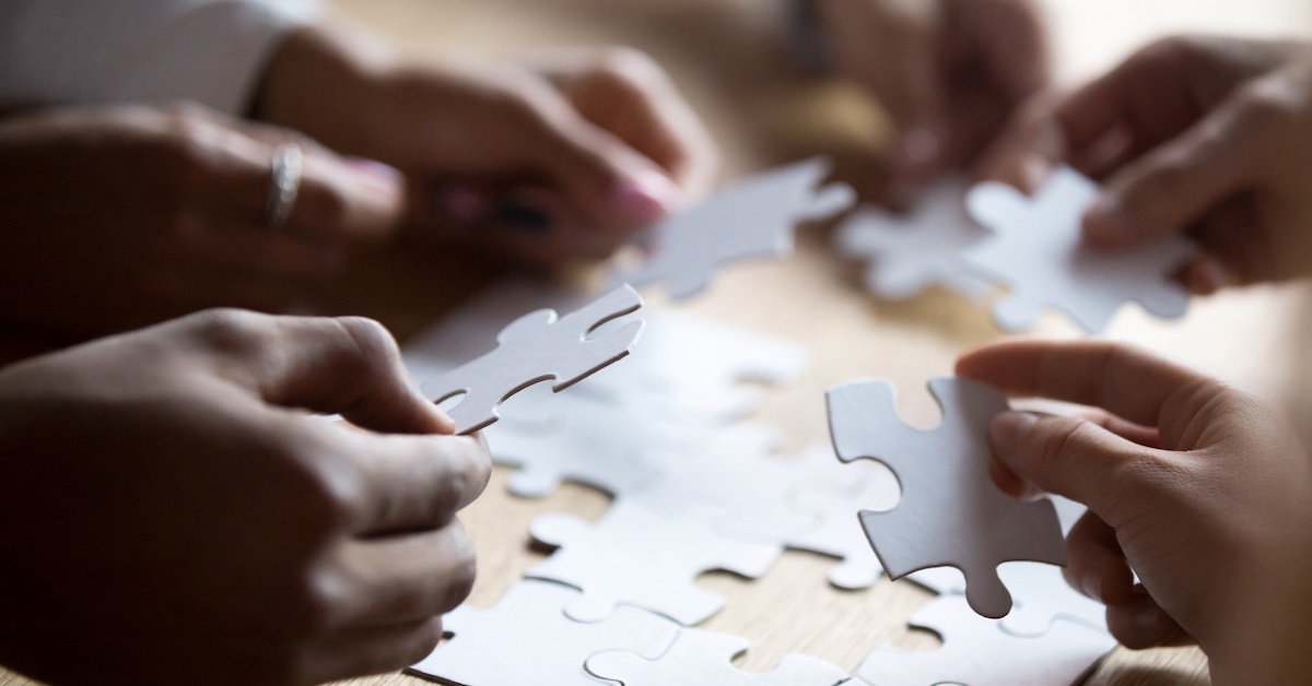 Close up of a table where at least six people's hands are putting together puzzle pieces.